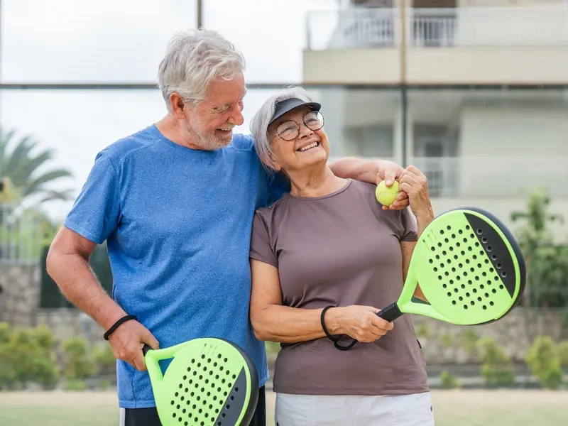 A smiling silver-haired couple with padel rackets, his arm around her shoulders