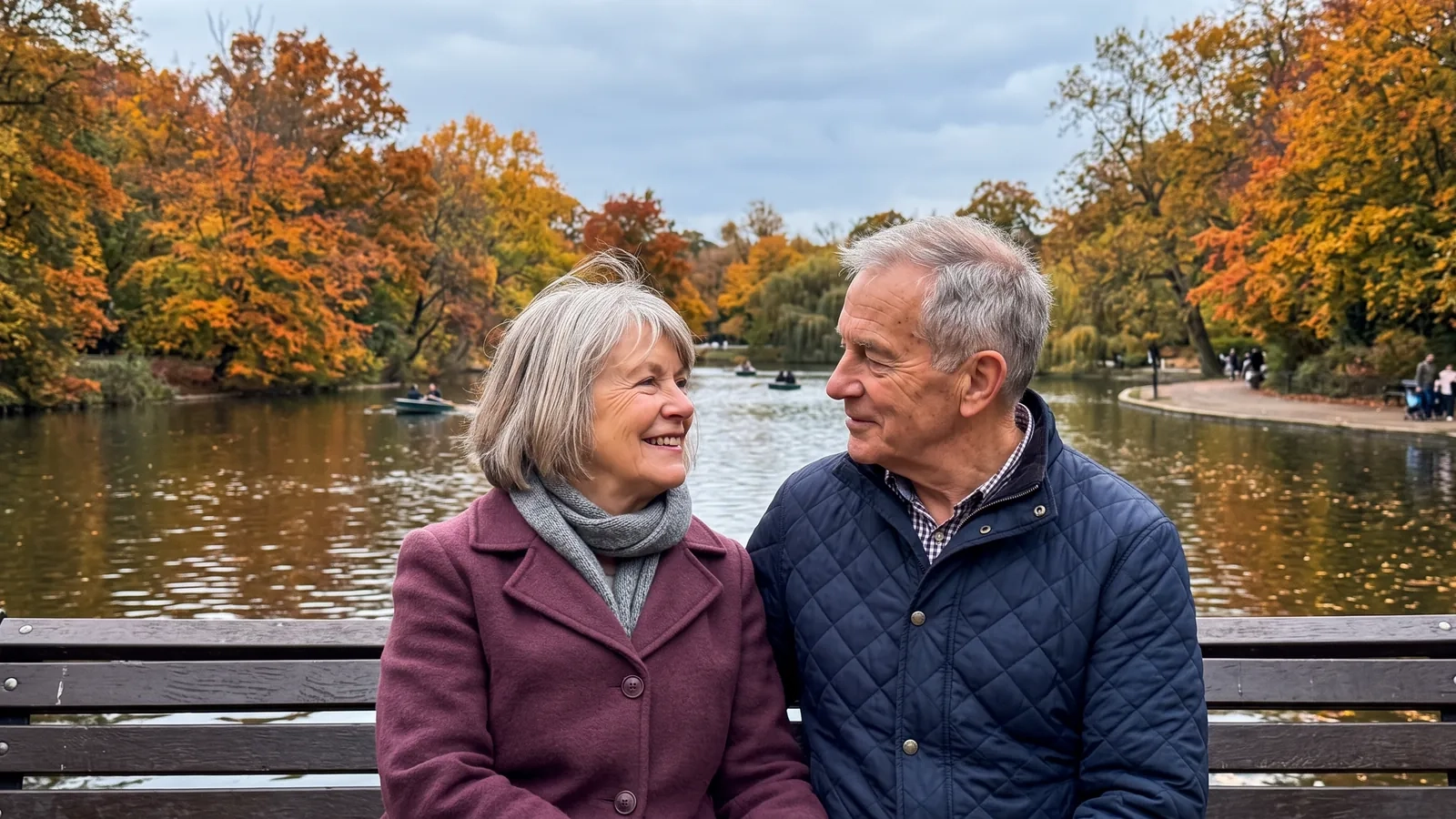 A couple in their late sixties sit close together on a bench beside a boating lake in a large park with orange and yellow autumn trees, turning to smile at each other (AI-generated image)