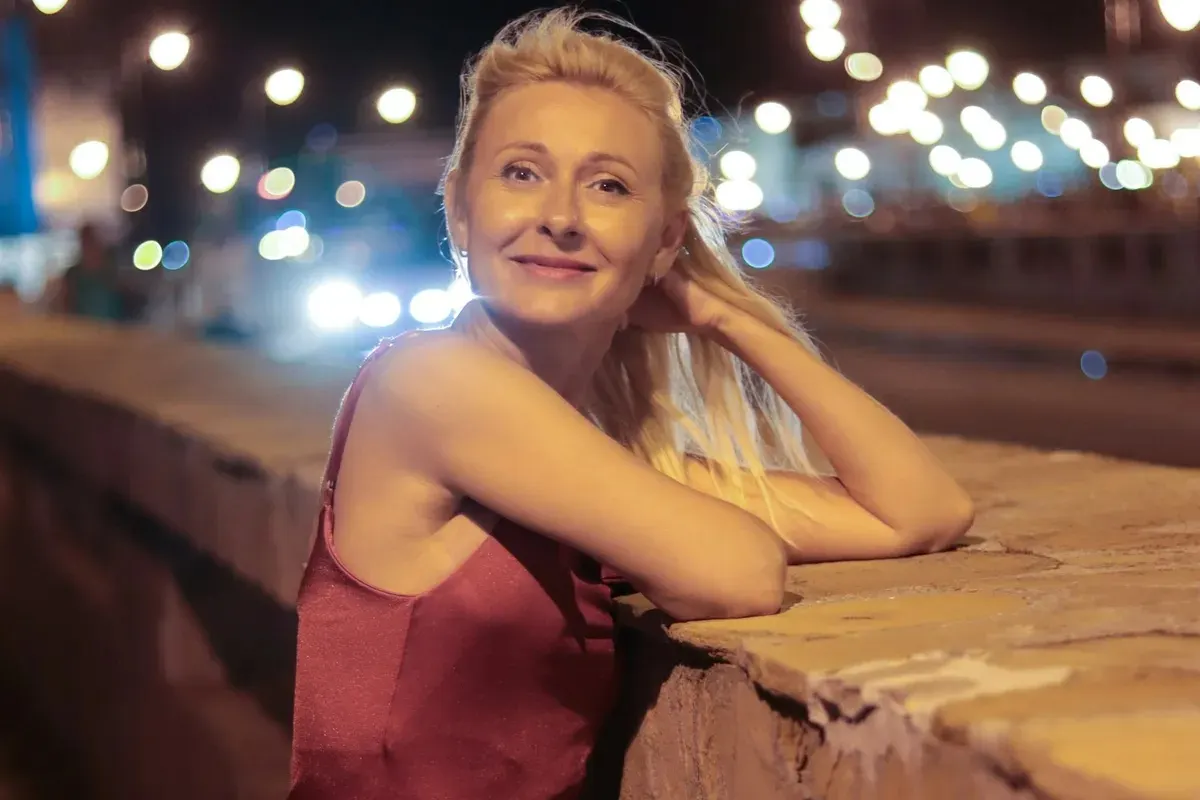 A blonde woman in a wine-red satin slip dress leaning on a wall at night, city lights behind her, smiling at the camera
