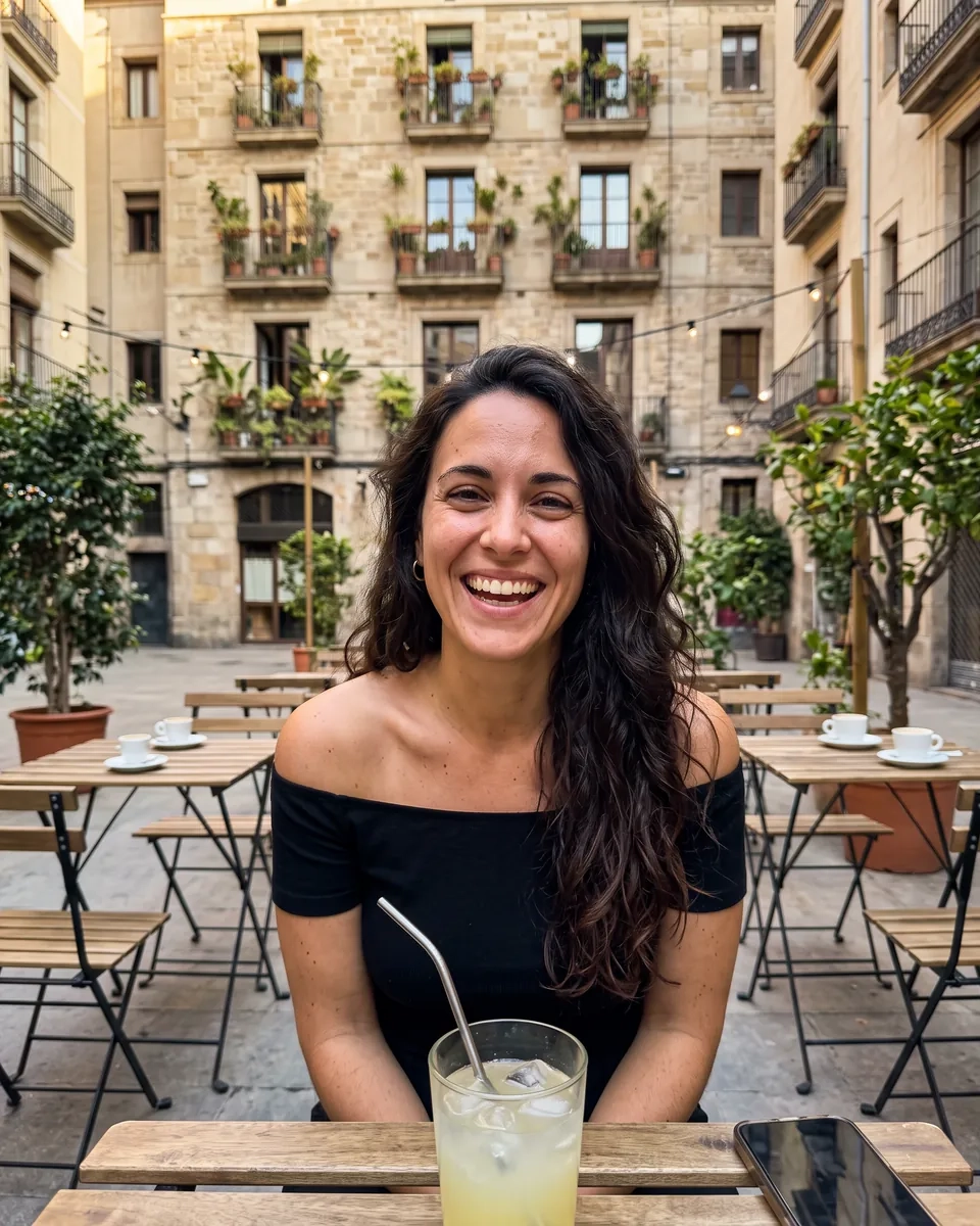 Una mujer de pelo largo oscuro con top negro de hombros descubiertos se ríe en una terraza de una plaza de Gràcia, en Barcelona, con una limonada en la mesa (imagen generada por IA)