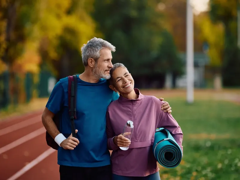 A smiling middle-aged couple with a yoga mat and water bottle at an outdoor running track