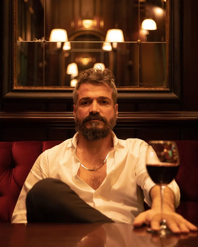 A bearded man in an open-collared white shirt sitting at a table in a warmly lit bar with a glass of red wine, looking straight at the camera