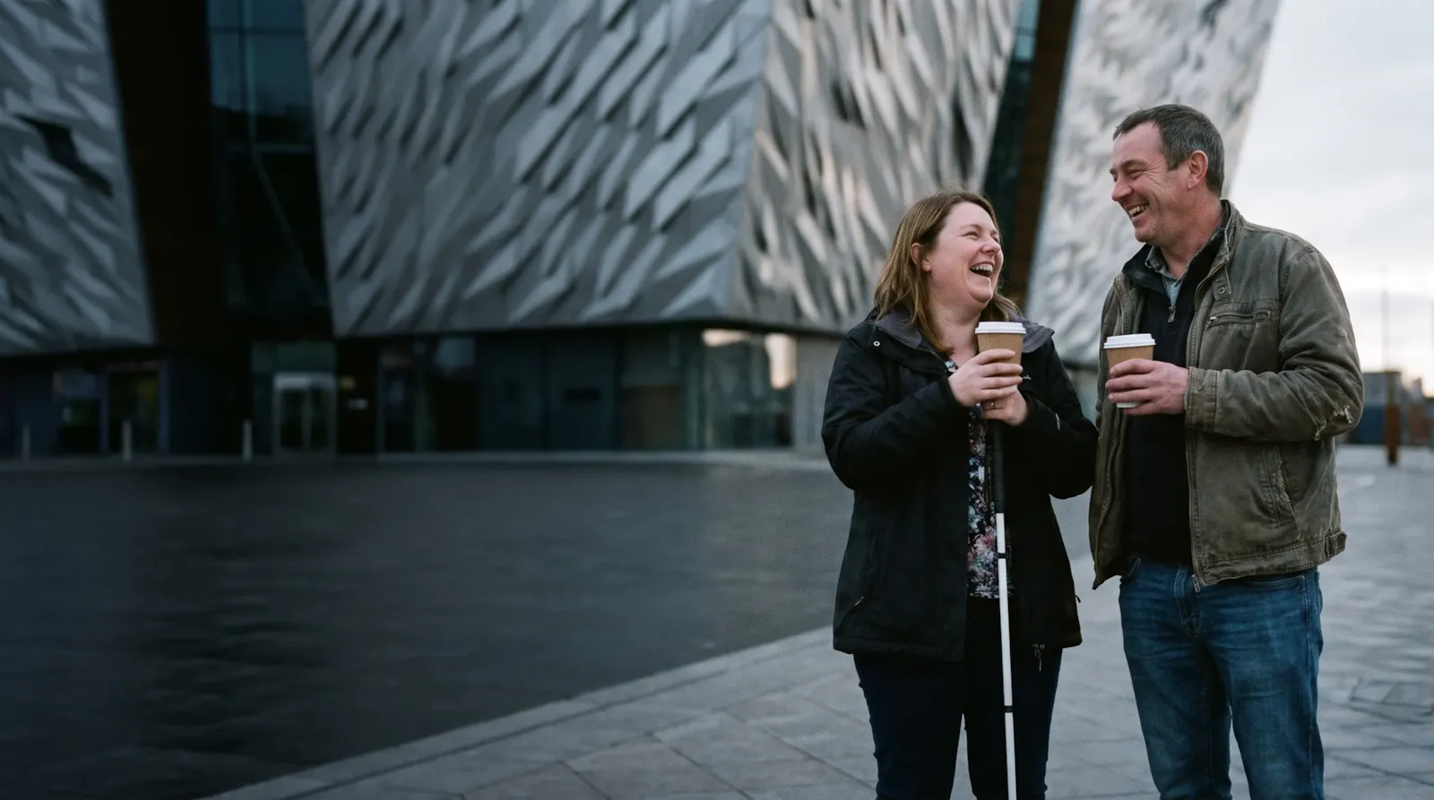 A couple laughing over coffee outside Titanic Belfast at dusk; the woman uses a white cane