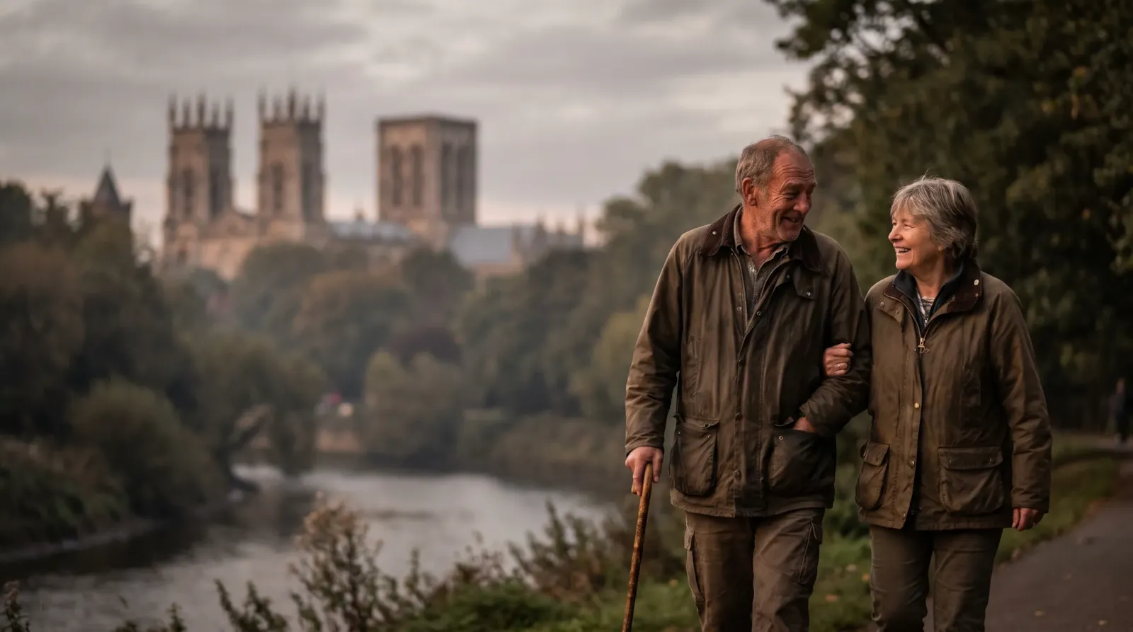 A couple arm in arm by the River Ouse in York with the Minster behind them; the man walks with a stick