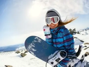 A smiling woman snowboarder in goggles holding her board on a sunny slope