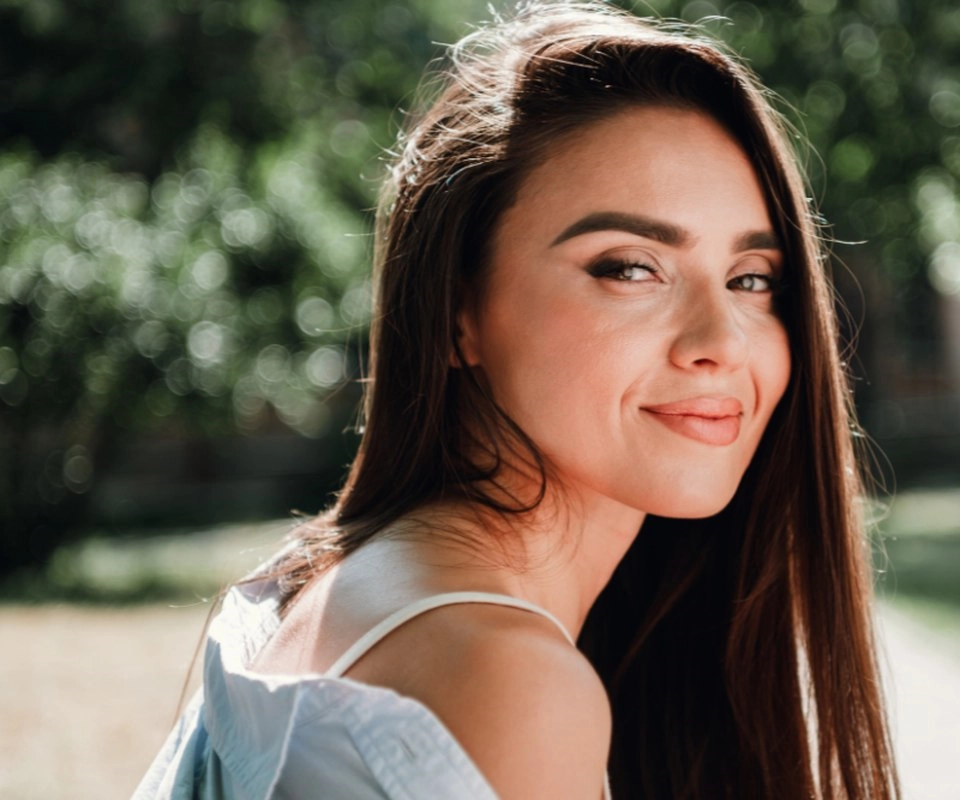 A woman with long dark hair smiles at the camera in the sunshine