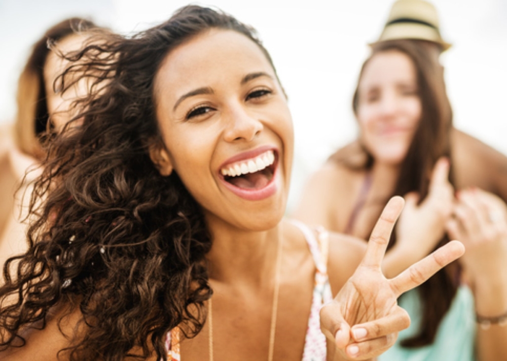 A woman with curly hair laughs and flashes a peace sign at the beach, with a friend in a straw hat behind her