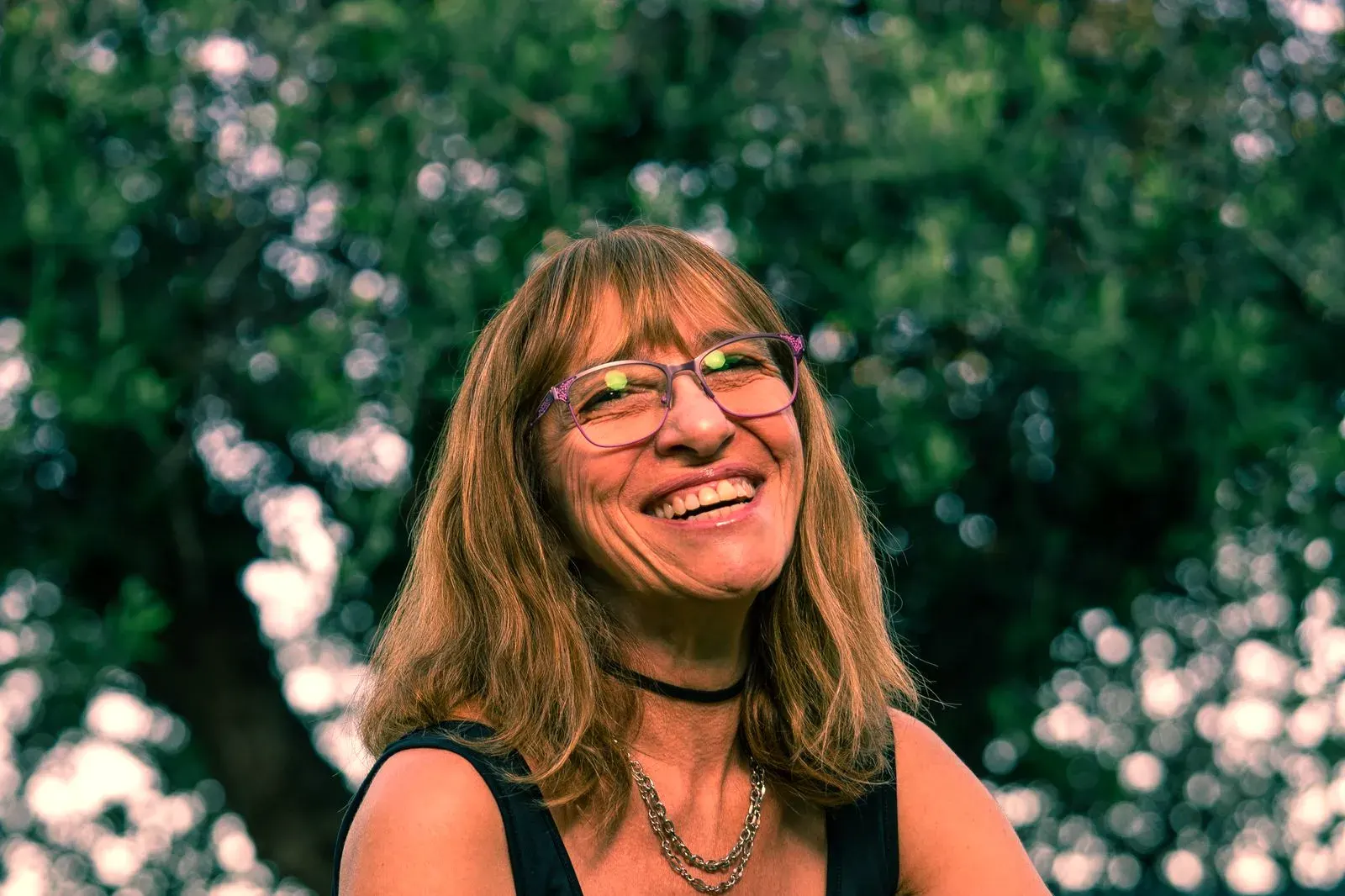 A laughing woman with shoulder-length hair and glasses in a sunny garden