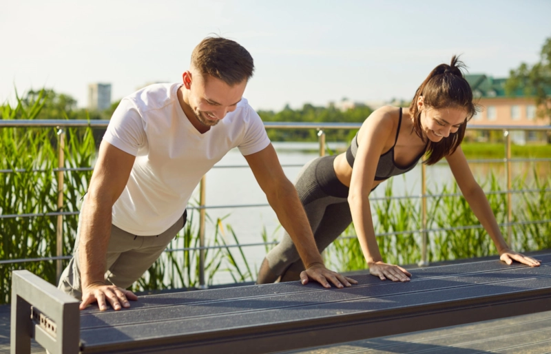 A smiling man and woman doing incline push-ups on a bench by a lake