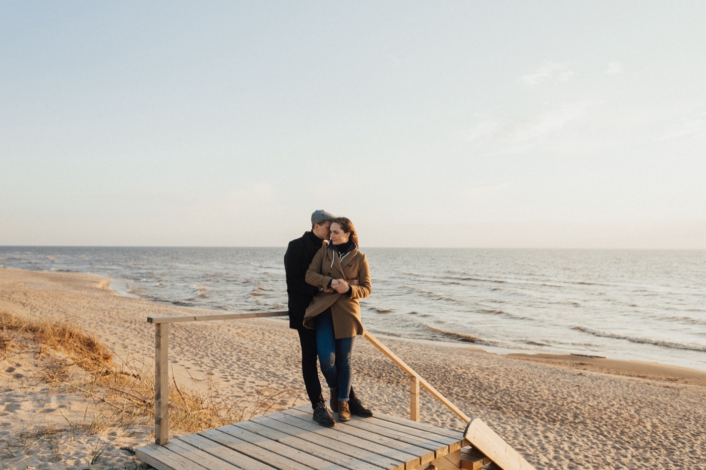 A man in a dark coat and flat cap embraces a woman in a tan coat on a wooden beach boardwalk
