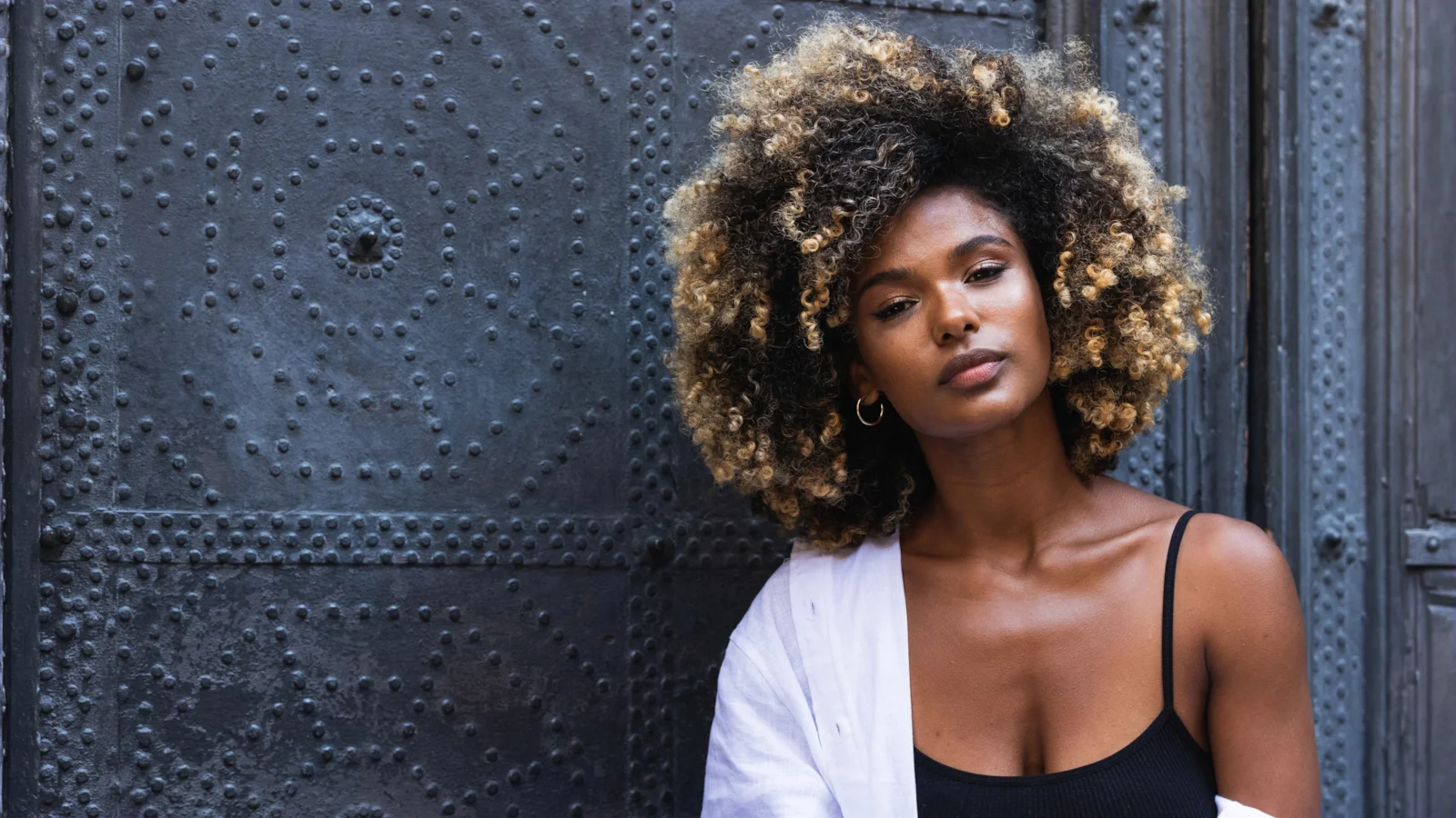 A woman with curly golden-tipped hair leaning against a dark studded door