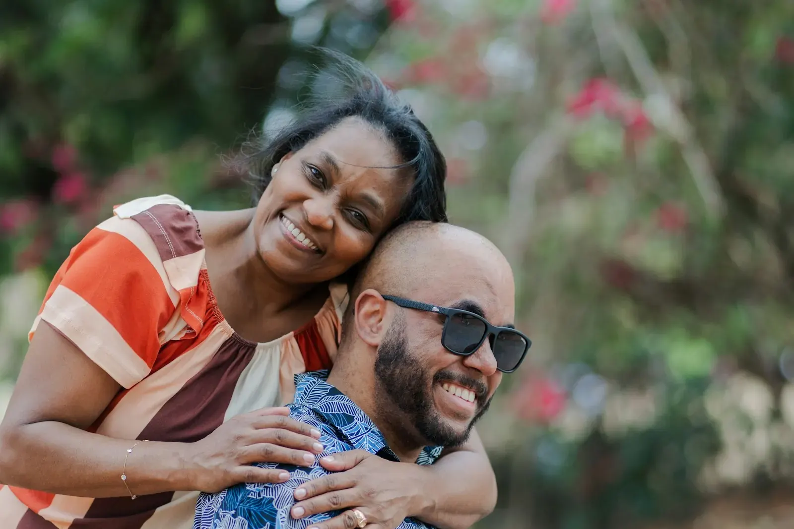 A laughing woman with her arms around a smiling man in sunglasses in a leafy park