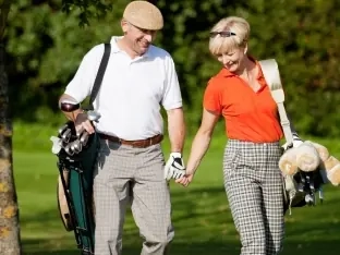 An older couple holding hands as they walk the course carrying their golf bags