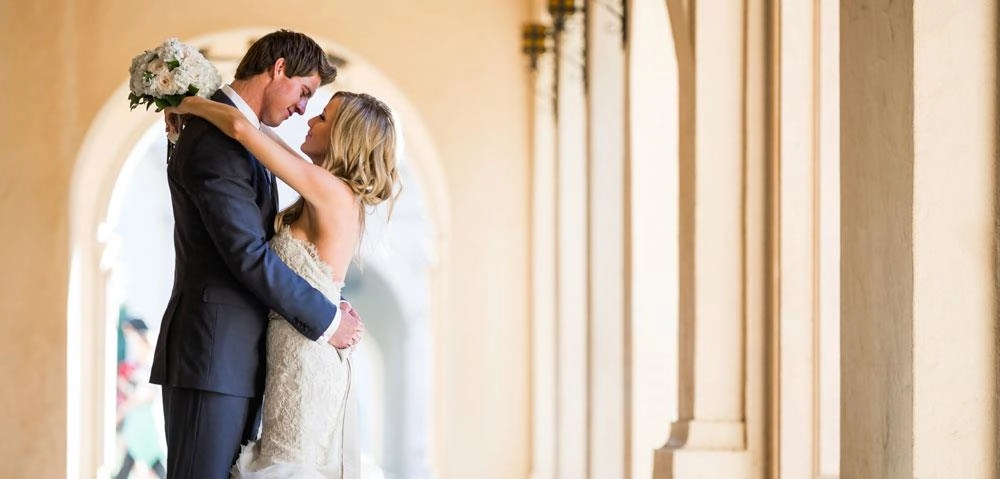 A bride and groom embracing beneath a row of sunlit arches