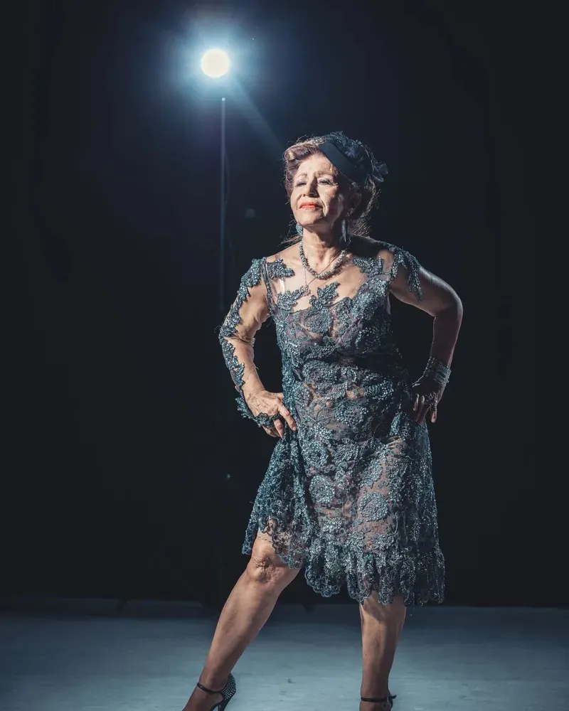 A woman with short auburn hair in a lace dress striking a confident pose under a single spotlight on a dark stage