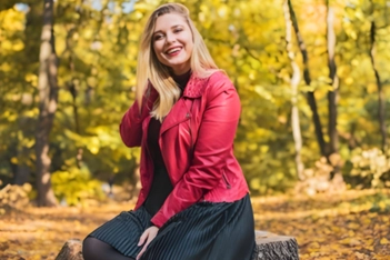 A laughing blonde woman in a red leather jacket sitting in an autumn wood
