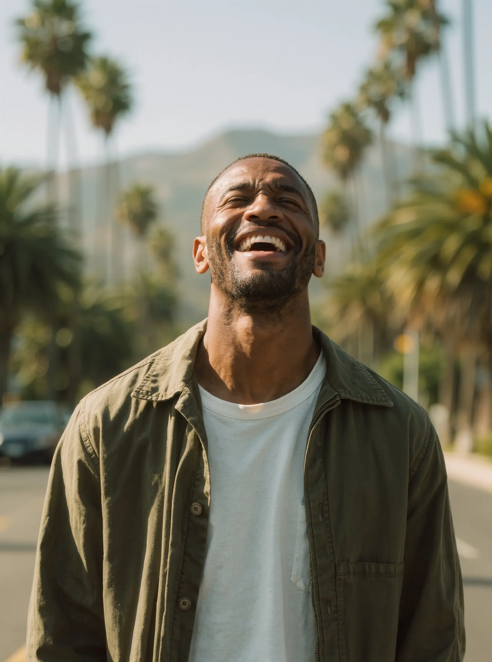 A man laughing on a sunny street lined with palm trees, hills in the distance