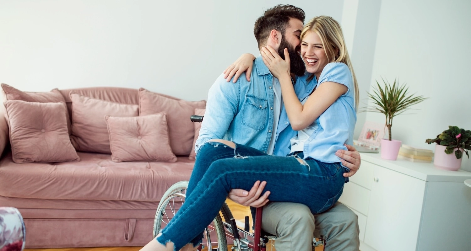 A man in a wheelchair and a woman sitting on his lap with her arm around his neck, both laughing together in a cozy living room