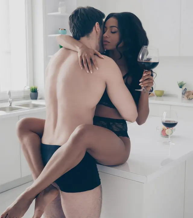 A man and a woman in black lingerie sharing a kiss in a bright kitchen with a glass of red wine, interracial hookups in Houston