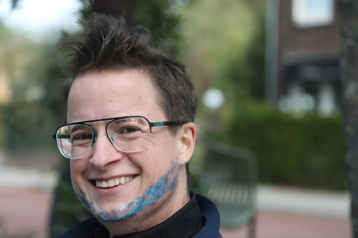 A smiling person with spiky dark hair, glasses and a glitter-dusted beard, photographed outdoors