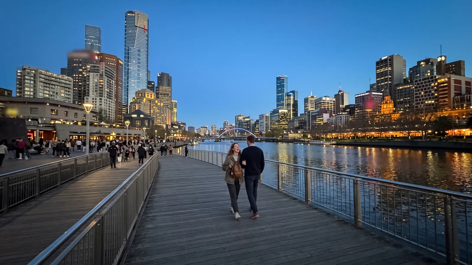 A couple walks arm in arm along the riverside promenade in Melbourne at dusk, the lit towers of the city reflected in the Yarra (AI-generated image)