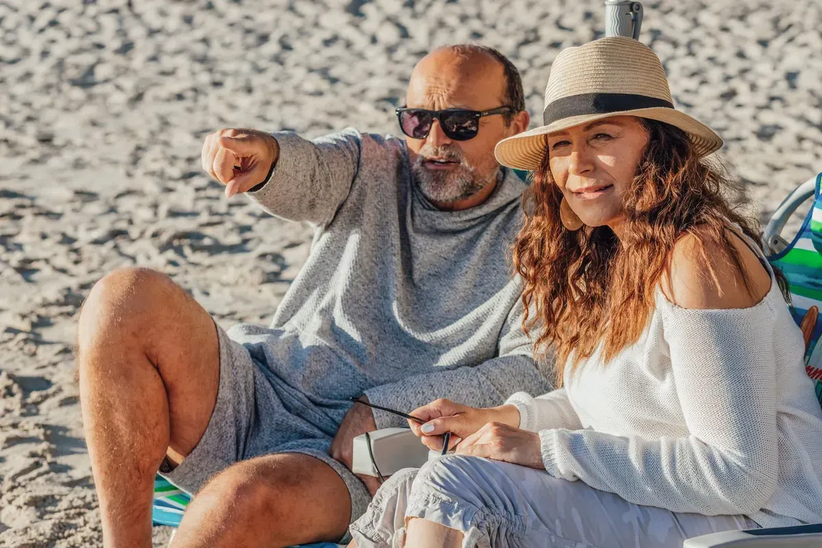 A laughing couple sitting on the sand, the man pointing at something along the beach