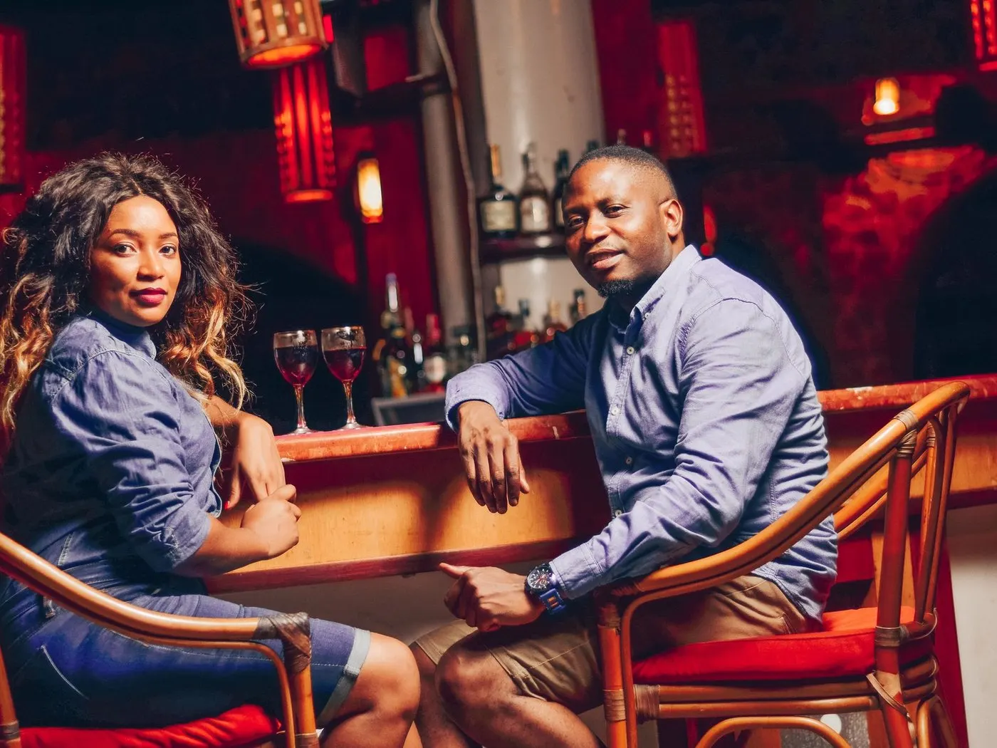 A man and a woman sitting side by side at a red-lit cocktail bar, two glasses of red wine between them, both looking at the camera