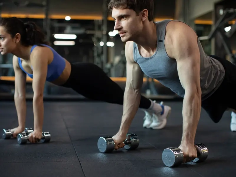 A man and woman doing dumbbell plank rows side by side in a gym