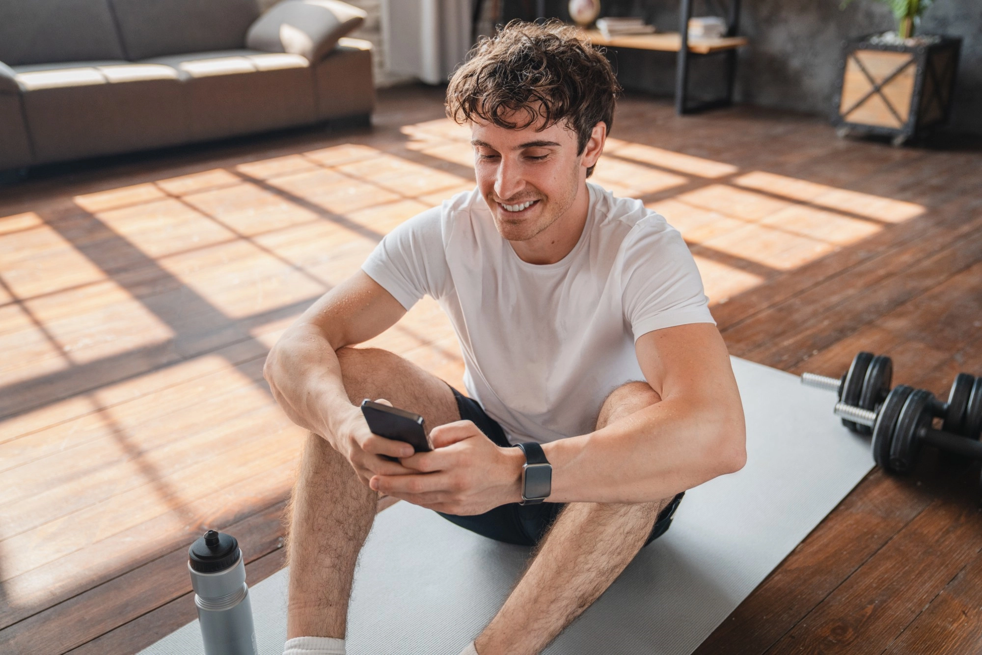 A smiling man sitting on a yoga mat at home checking his phone after a workout