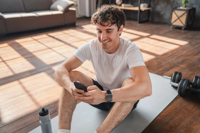 A smiling man sitting on a yoga mat at home checking his phone after a workout