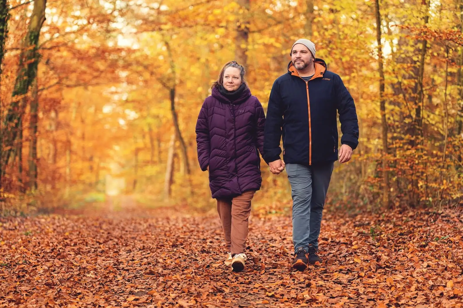 A couple in warm jackets walking hand in hand along a leaf-covered woodland path in autumn light