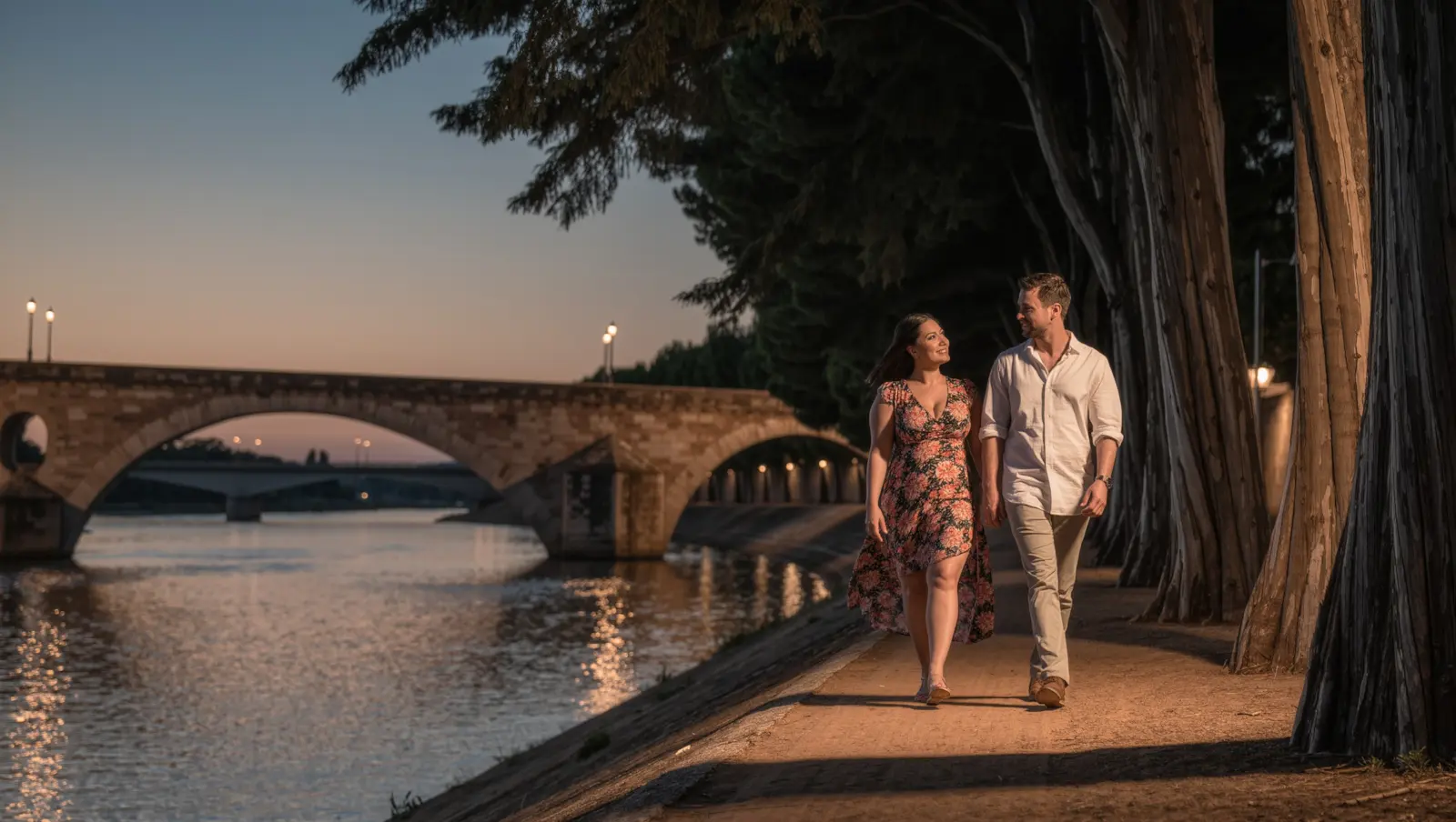 A woman in a floral dress and a man in a pale shirt walking along a riverside path beside a stone arch bridge at dusk