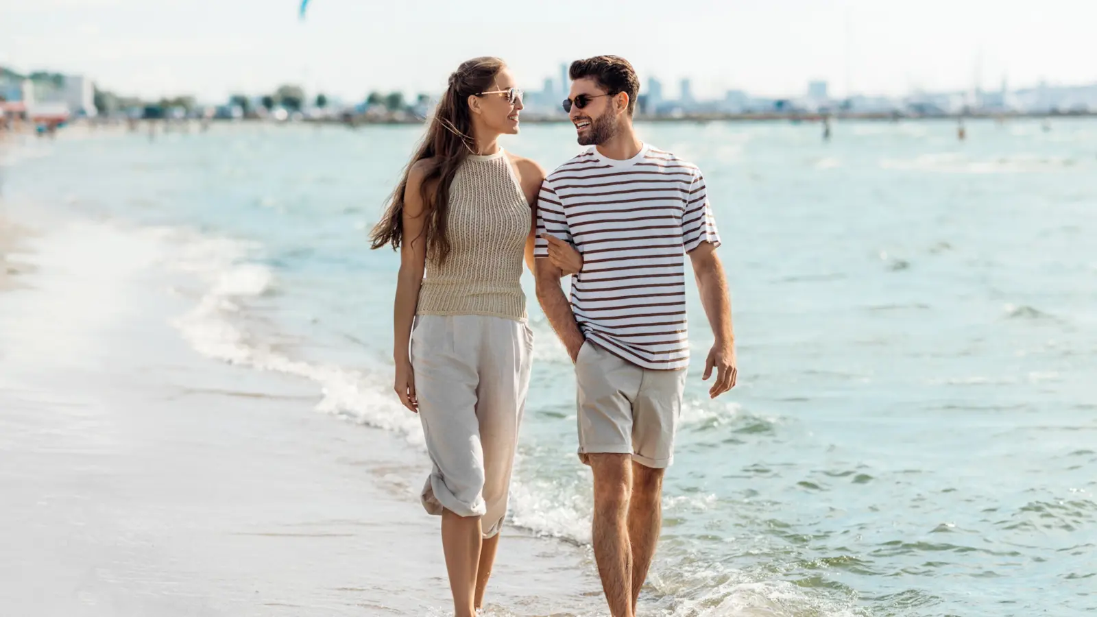 A woman and man walking together along a sunny beach, talking and smiling