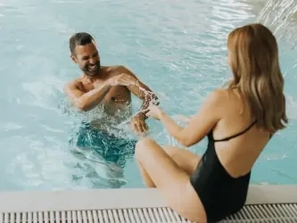 A laughing man in the pool splashing a woman sitting on the pool edge