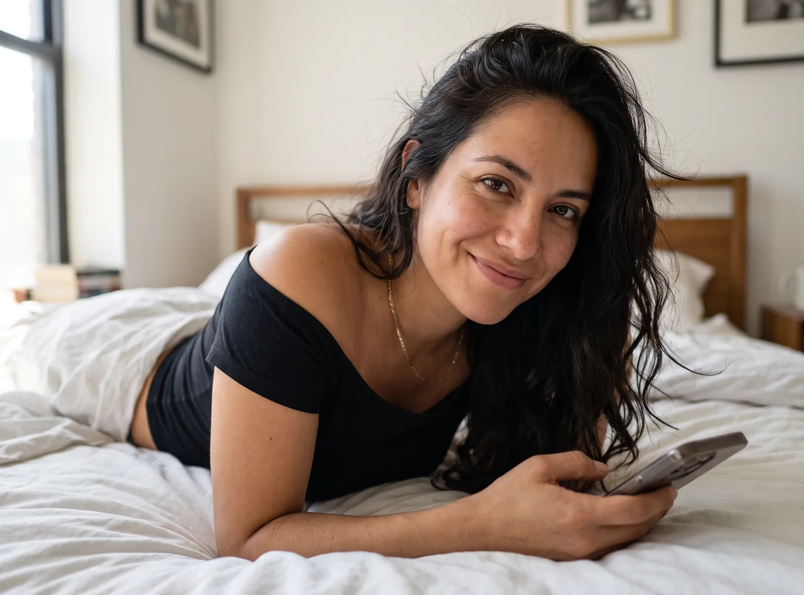 Una mujer de pelo largo y oscuro, tumbada de lado en una cama con sábanas blancas, mira a la cámara con una sonrisa cómplice mientras sostiene su móvil (imagen generada por IA)