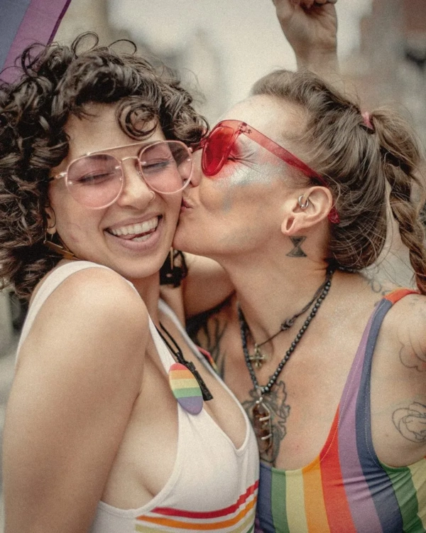 Two women in rainbow outfits sharing a kiss on the cheek at Pride