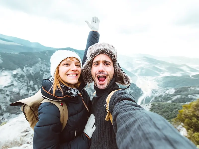 A laughing couple in winter hats taking a selfie on a snowy mountain lookout