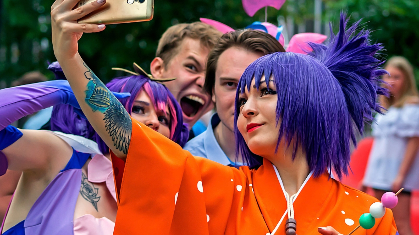 A cosplayer in a purple wig and orange costume taking a selfie with friends