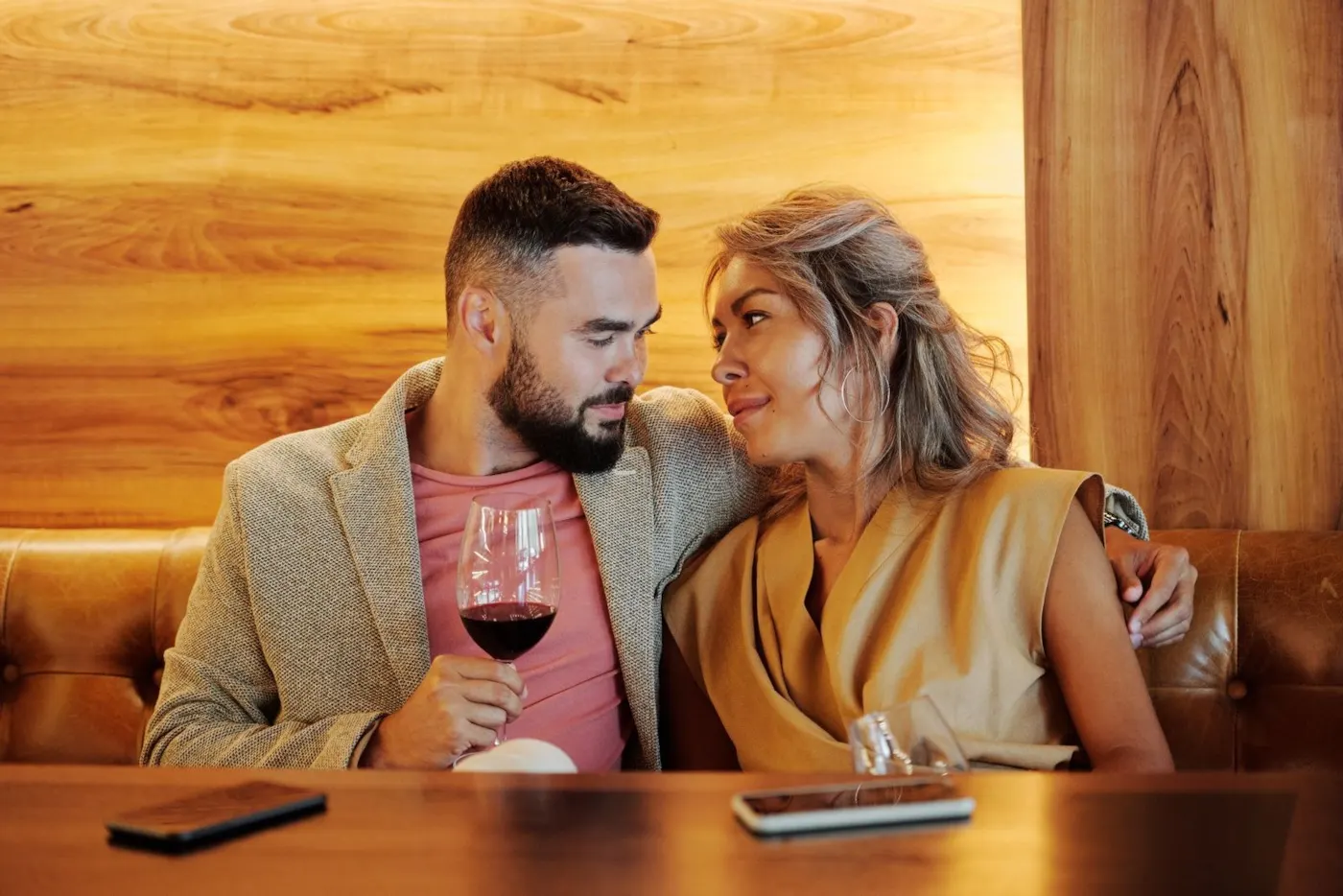 A man and a woman sitting close in a warmly lit wine bar booth, sharing red wine and a lingering look