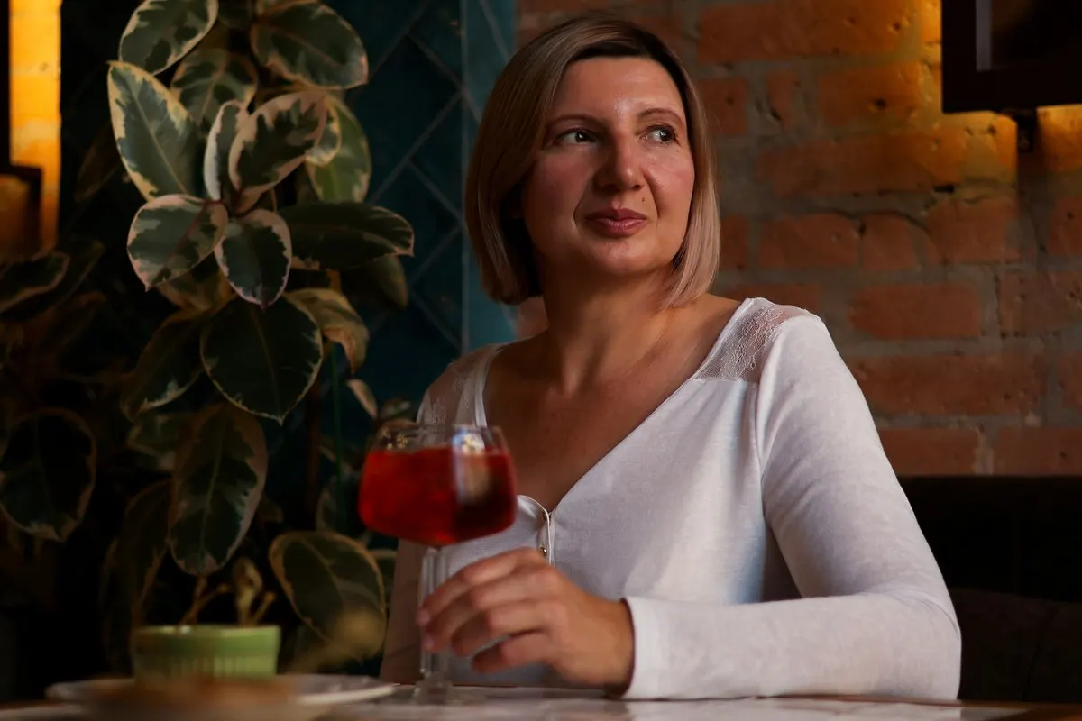 A woman in her forties in a white top with a glass of red cocktail, glancing across a candlelit bar with a brick wall behind her