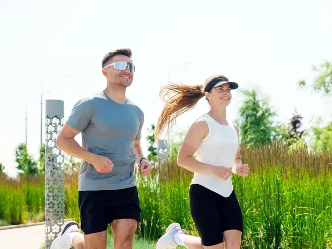 A man and woman running together along a sunny city path lined with tall grasses