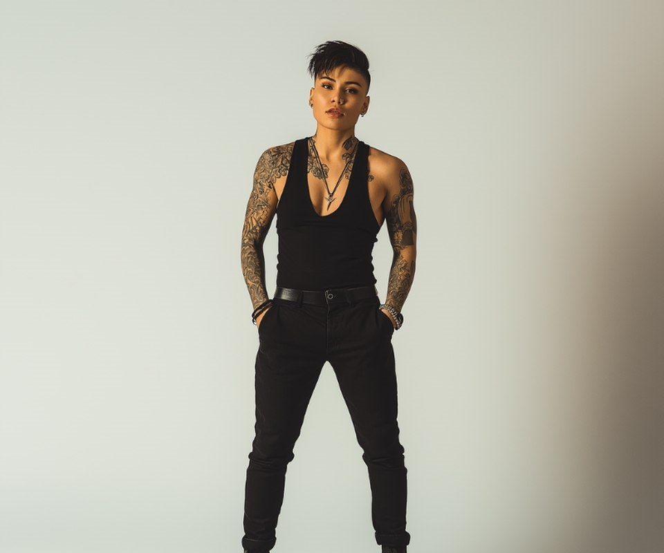 A butch woman standing with hands in pockets, tattooed arms, in a plain studio
