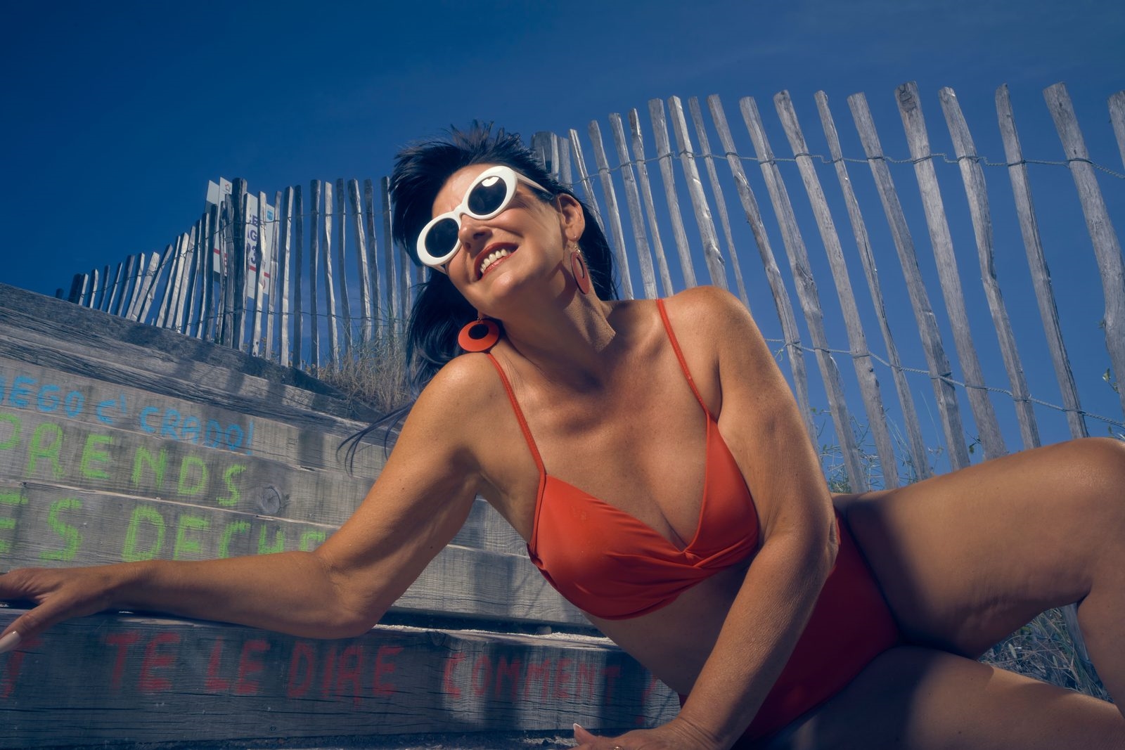 Smiling mature woman in a red bikini and white sunglasses lounging at the beach
