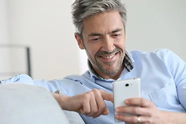 A man with grey hair smiling at his phone on a sofa