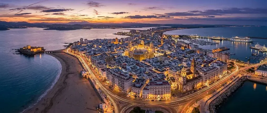 Vista aérea de Cádiz al anochecer, con la península del casco antiguo, la playa de La Caleta y el puerto iluminados