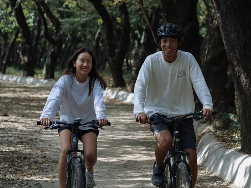A couple riding side by side and smiling on a tree-lined gravel path