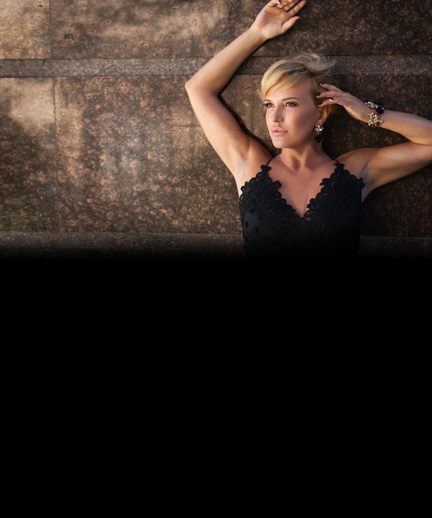 A blonde woman in a black lace dress leaning back against a sunlit stone wall, arms raised, looking away