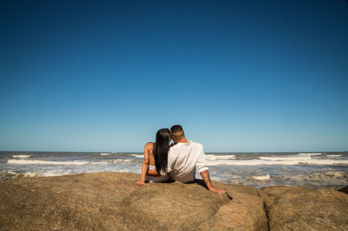 A man and woman sit side by side on rocks overlooking ocean waves under a clear blue sky