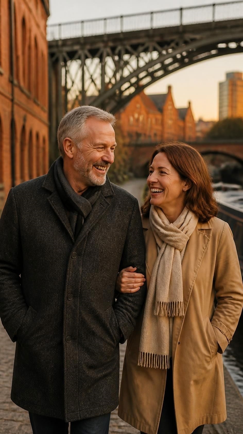 A happy couple walking arm in arm along a canal towpath with red brick warehouses and an iron railway viaduct behind them