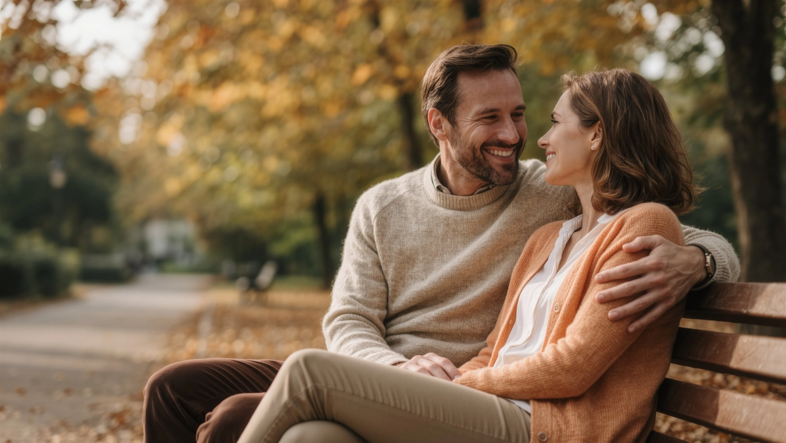 A smiling couple in their forties sitting close together on a park bench among autumn trees